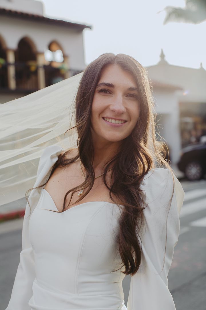 A bride in a white wedding dress stands outdoors with a flowing veil caught in the breeze.