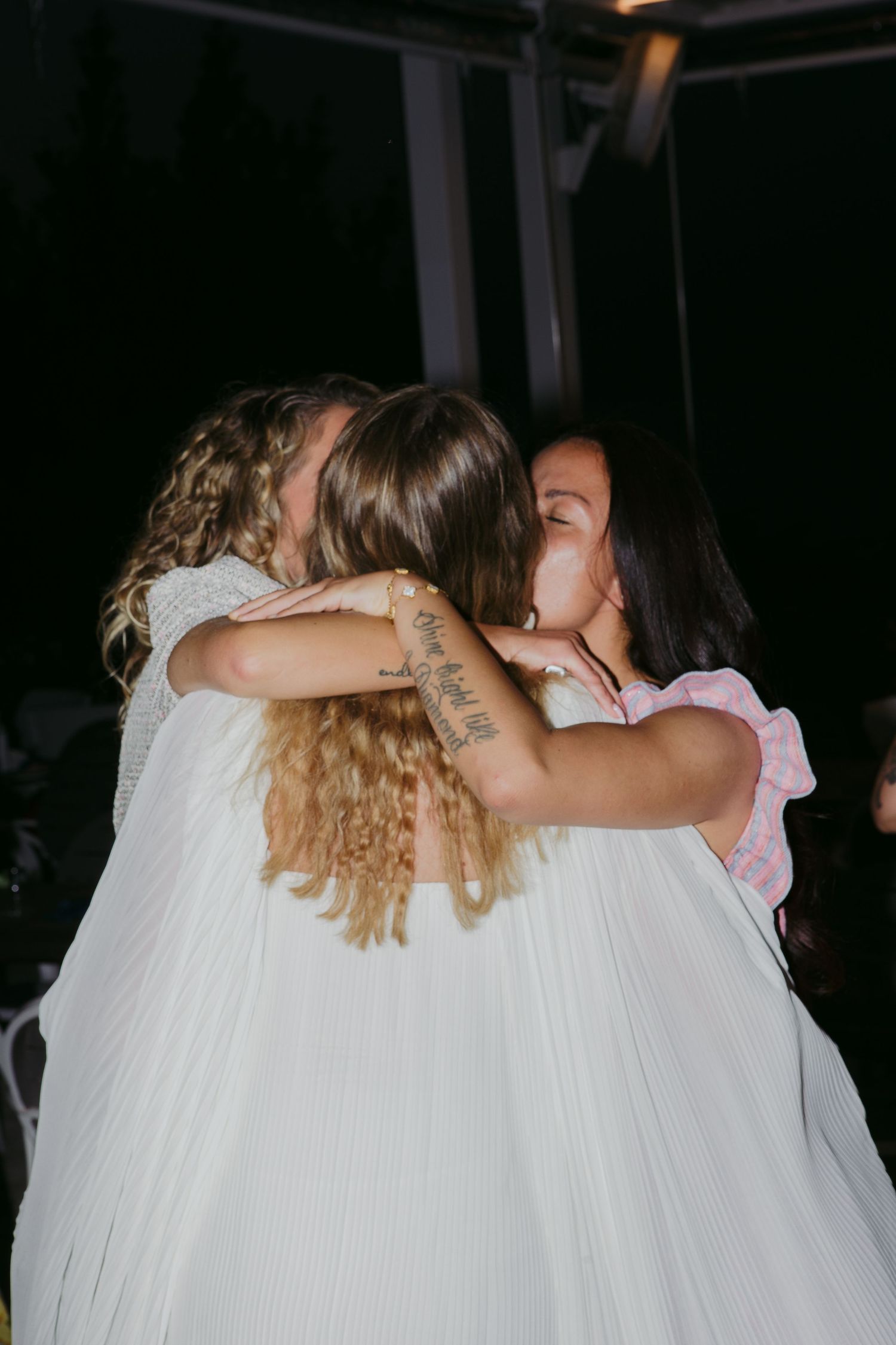Three people share an emotional embrace at what appears to be a wedding celebration in a dimly lit setting.
