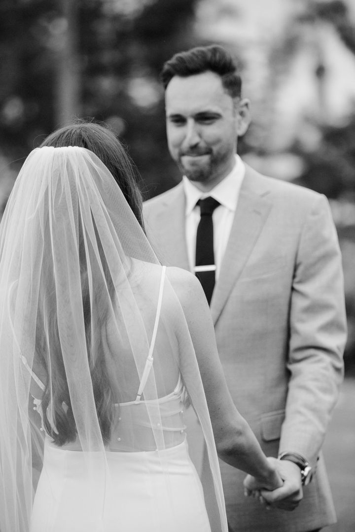 Black and white photo of a groom in a suit holding hands with a bride in a white gown and veil at a wedding.