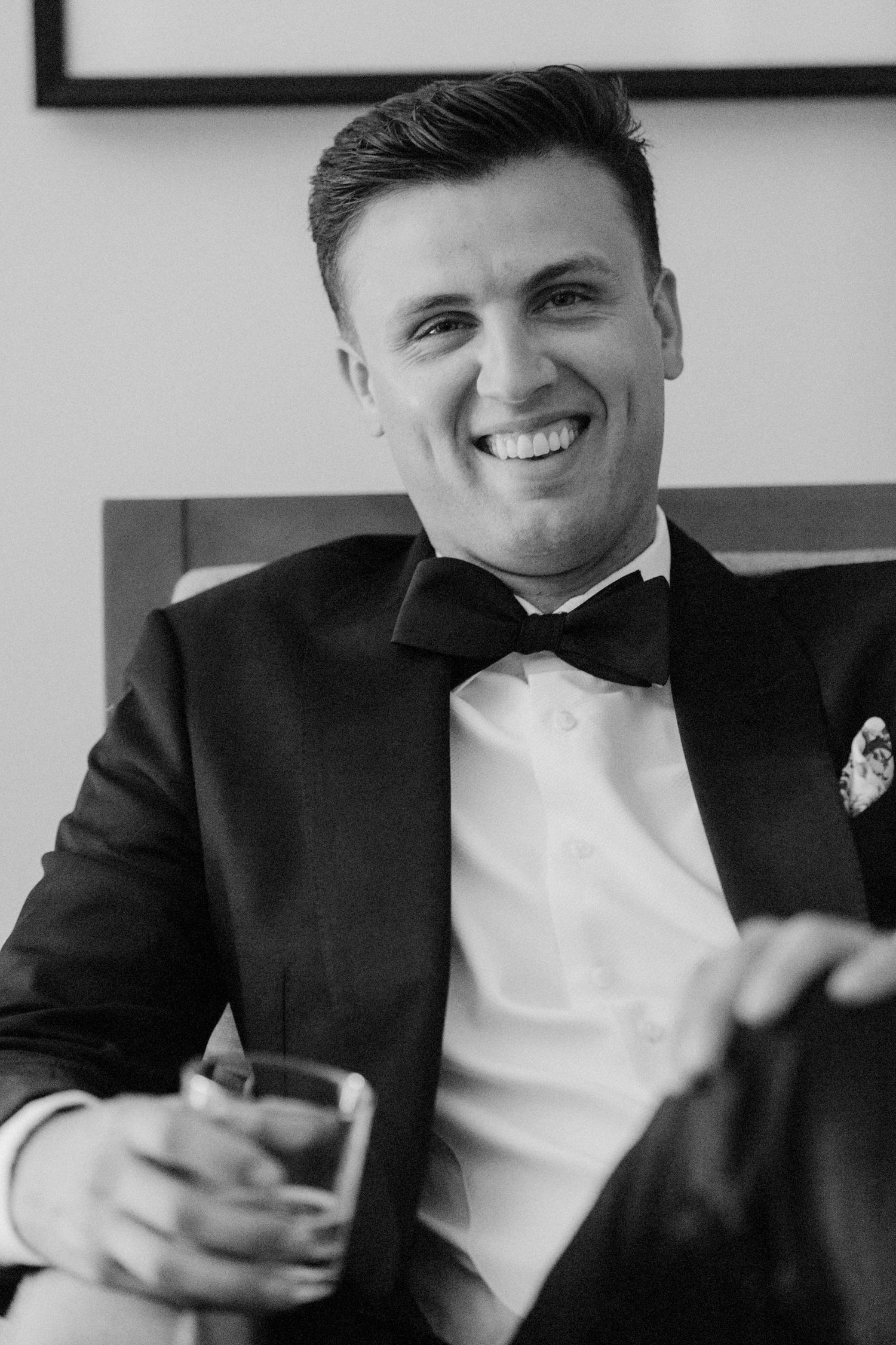Black and white portrait of person in tuxedo smiling while holding a drink at formal event.