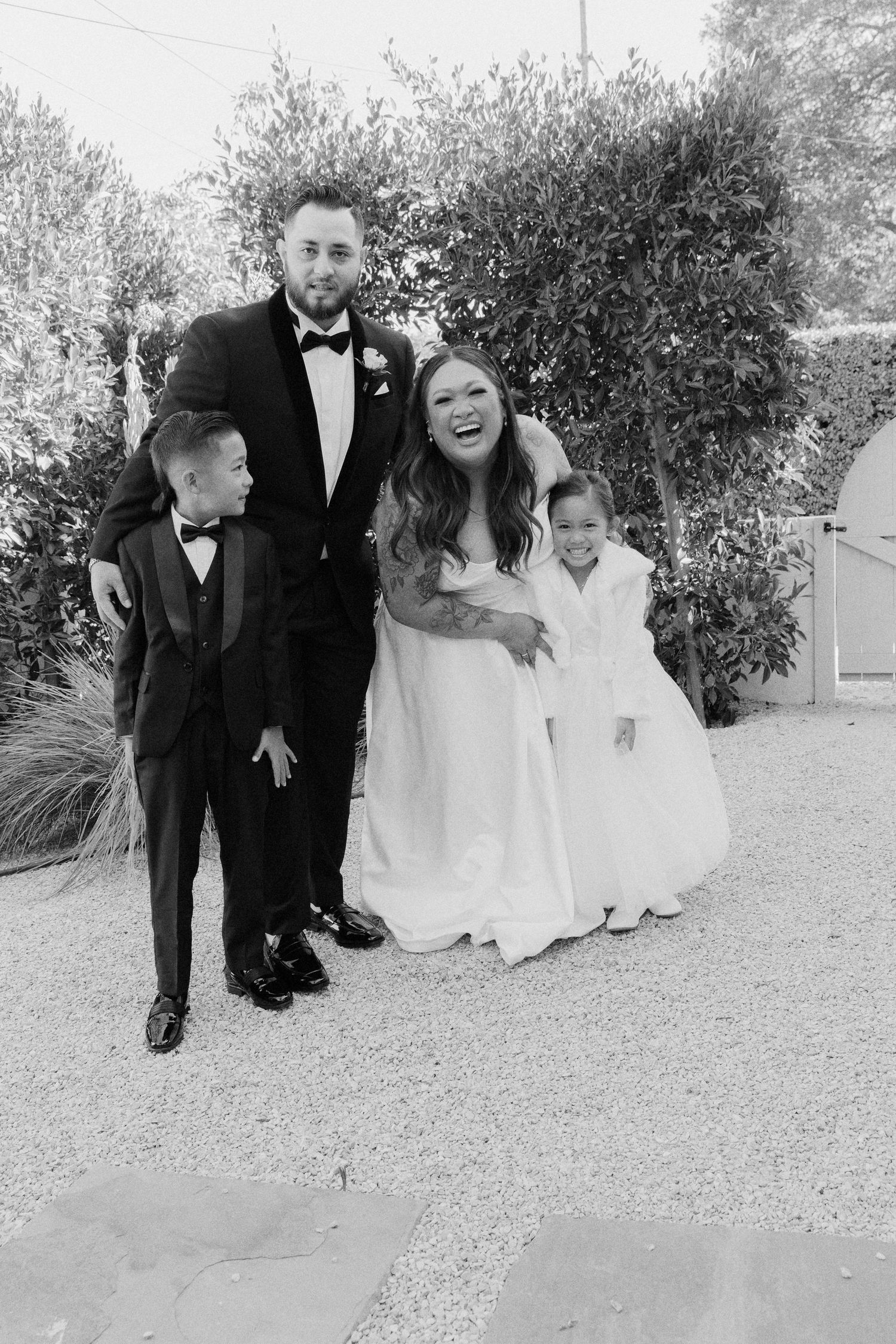 A joyful wedding party in formal attire poses together outdoors against greenery in black and white.