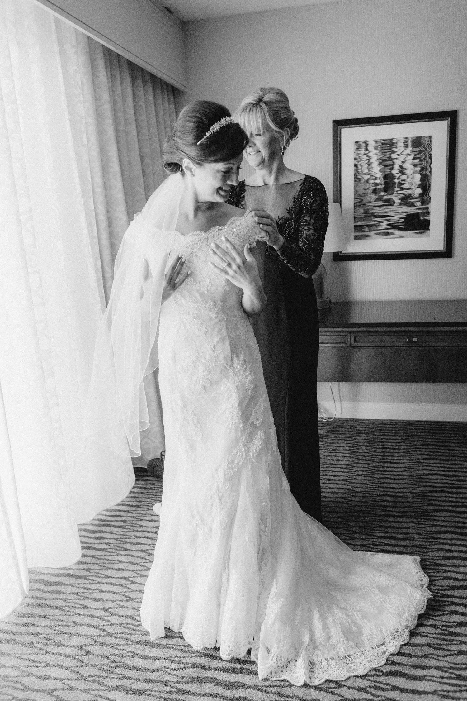 Black and white photo of a bride in lace gown and veil sharing a tender moment with her mother before the wedding.