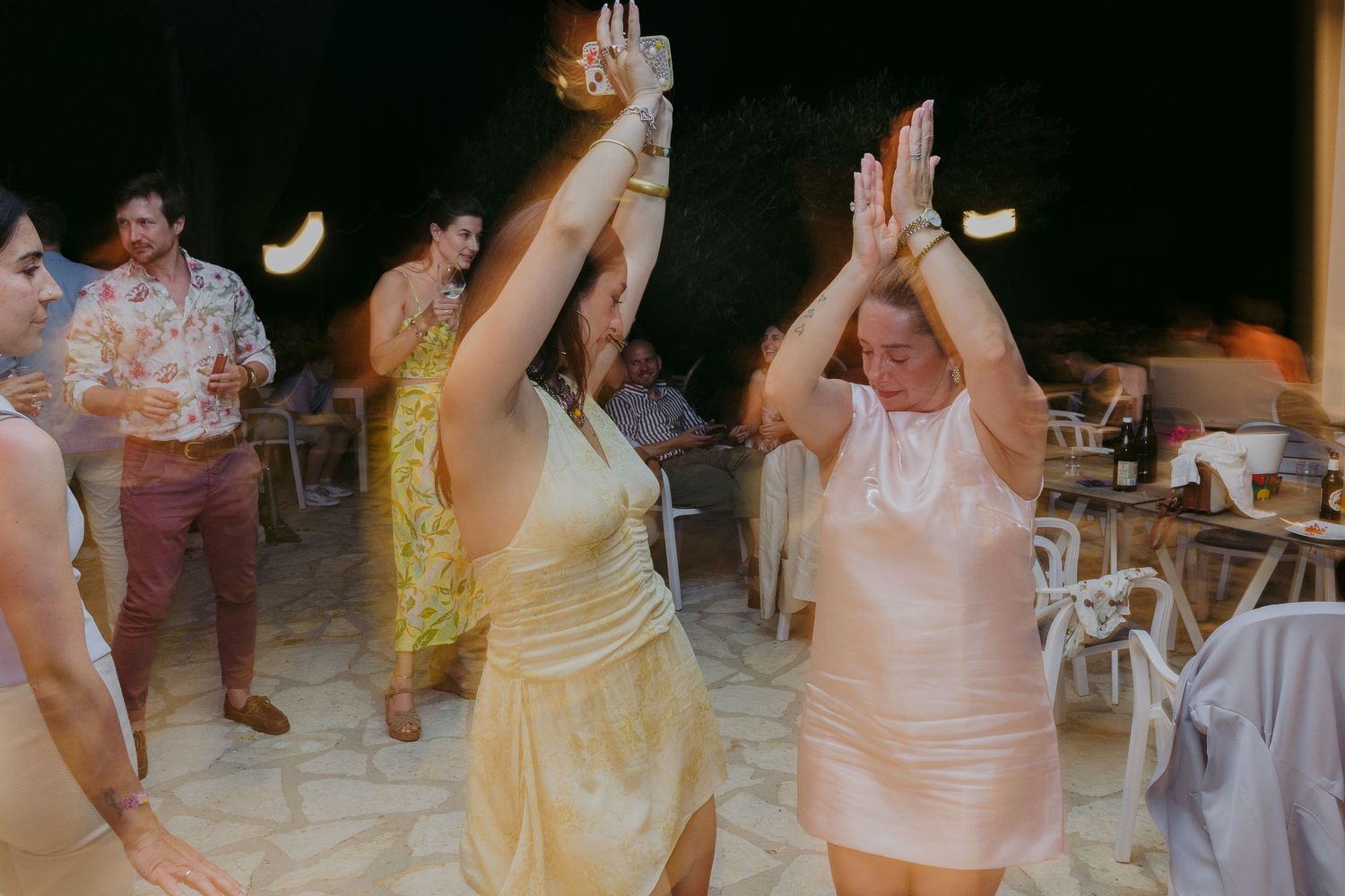 Two women dancing joyfully with raised hands at an outdoor wedding reception celebration at night.