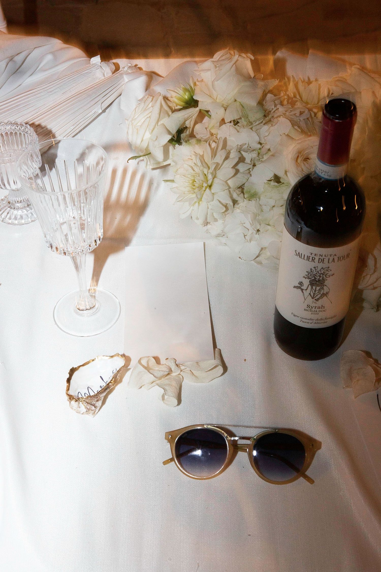 Wine bottles and sunglasses arranged on a white tablecloth with delicate floral decorations and crystal glassware.
