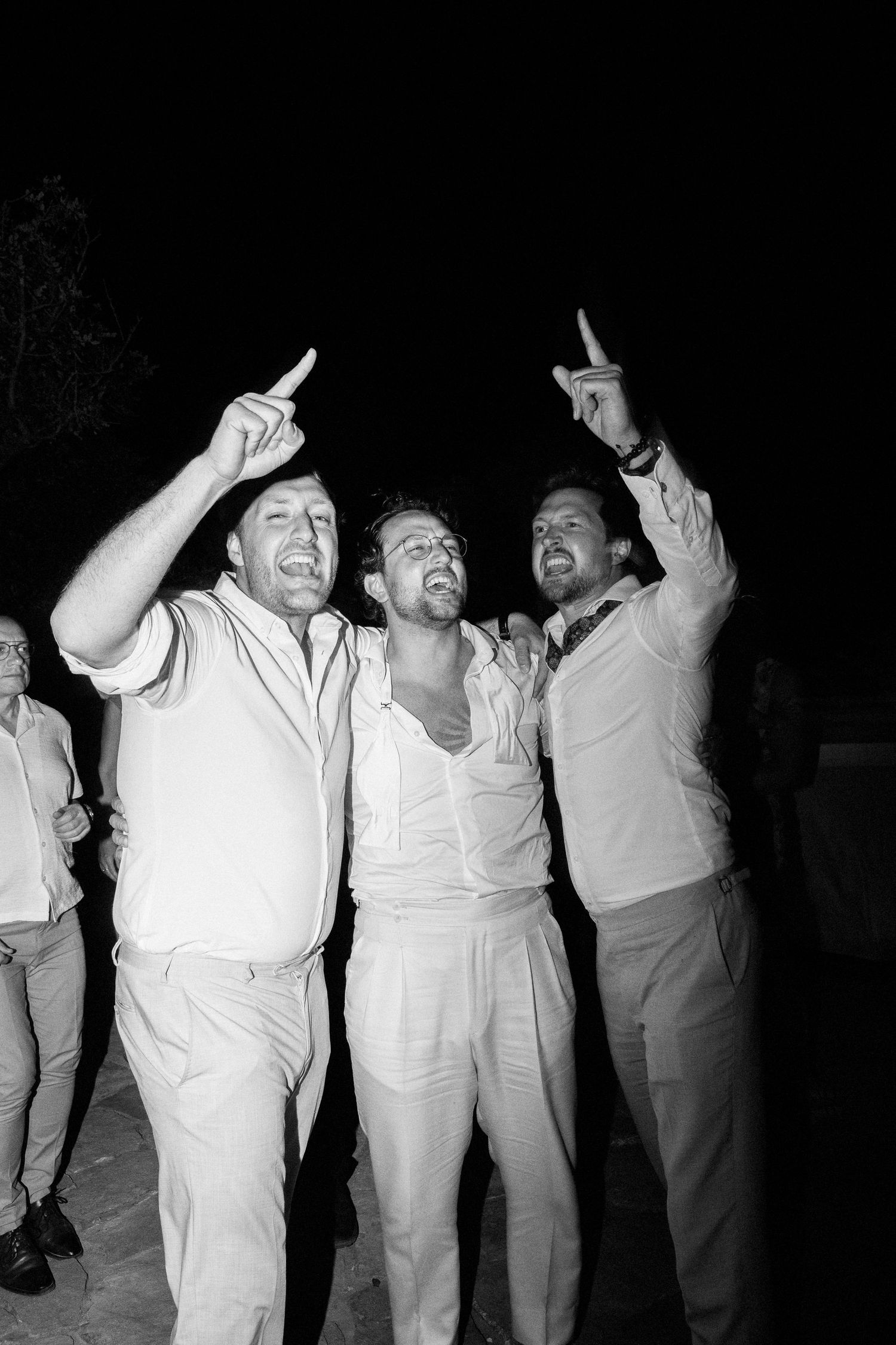 Three people in casual white and light-colored clothing point upward while celebrating together in a black and white photo.