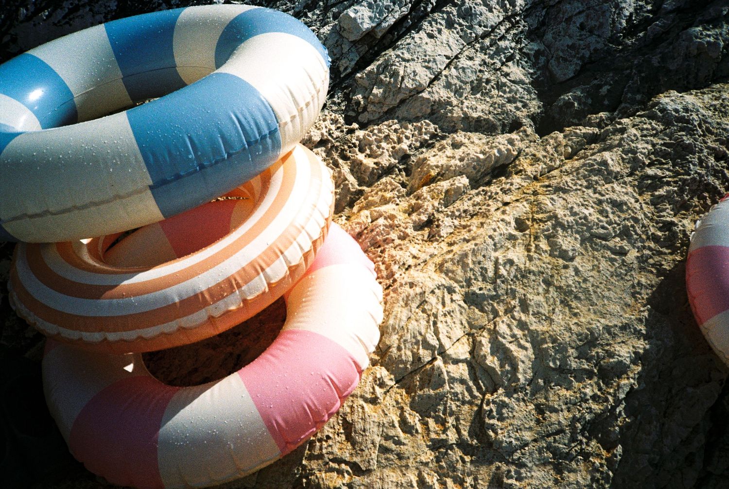 Colorful inflatable swim rings stacked on rocky ground in warm sunlight.