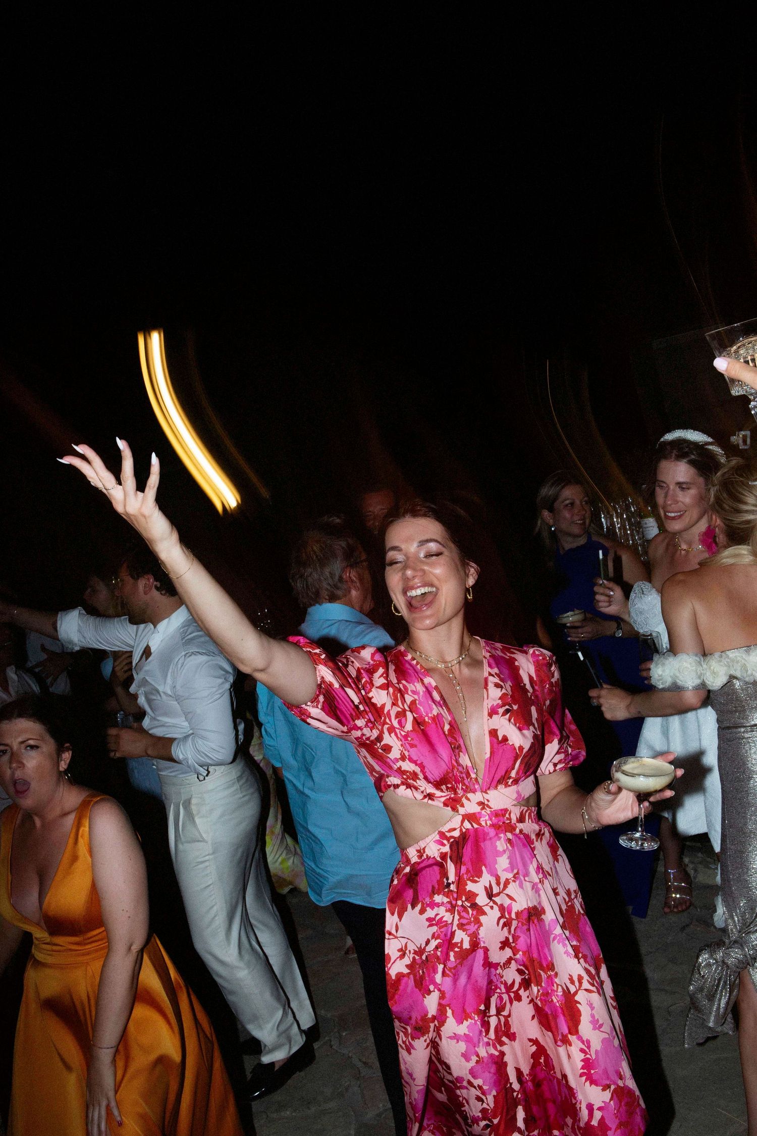 A person in a pink floral dress dances enthusiastically at a party with others visible in the background.