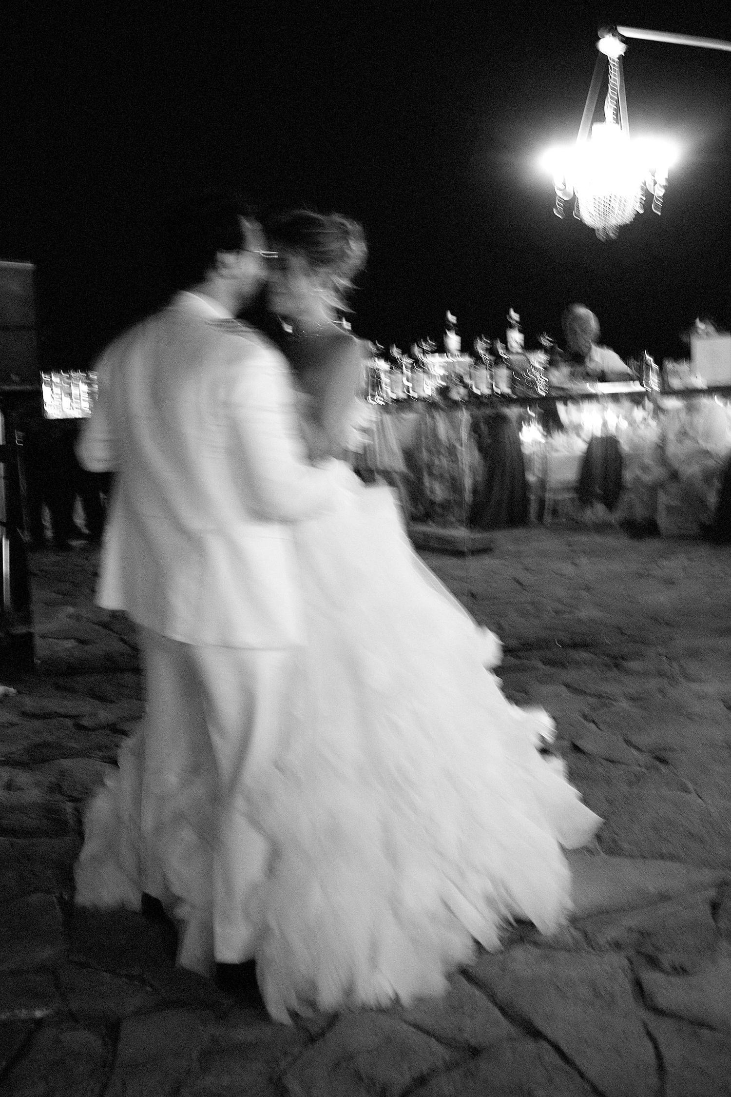 A blurred black and white photo of a couple dancing at a wedding reception under a crystal chandelier.