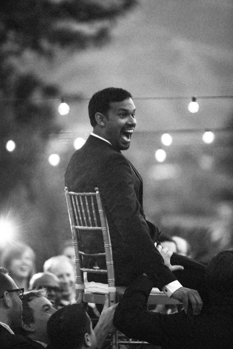 A person in a suit sits on a chair at an event, turning back with an animated expression while string lights glow above.