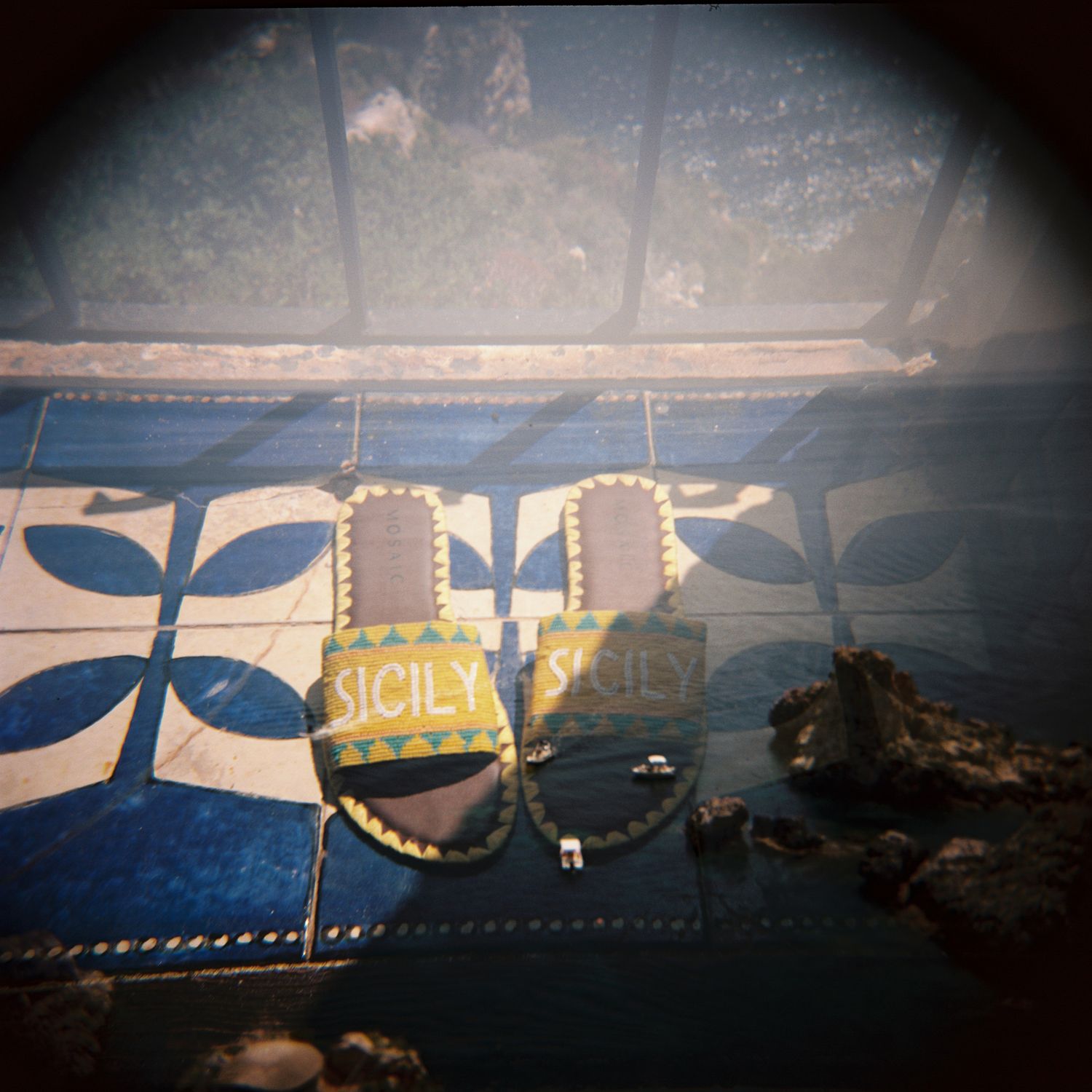 Colorful Sicily-branded sandals displayed on blue floral tiles with a scenic coastal view in the background.