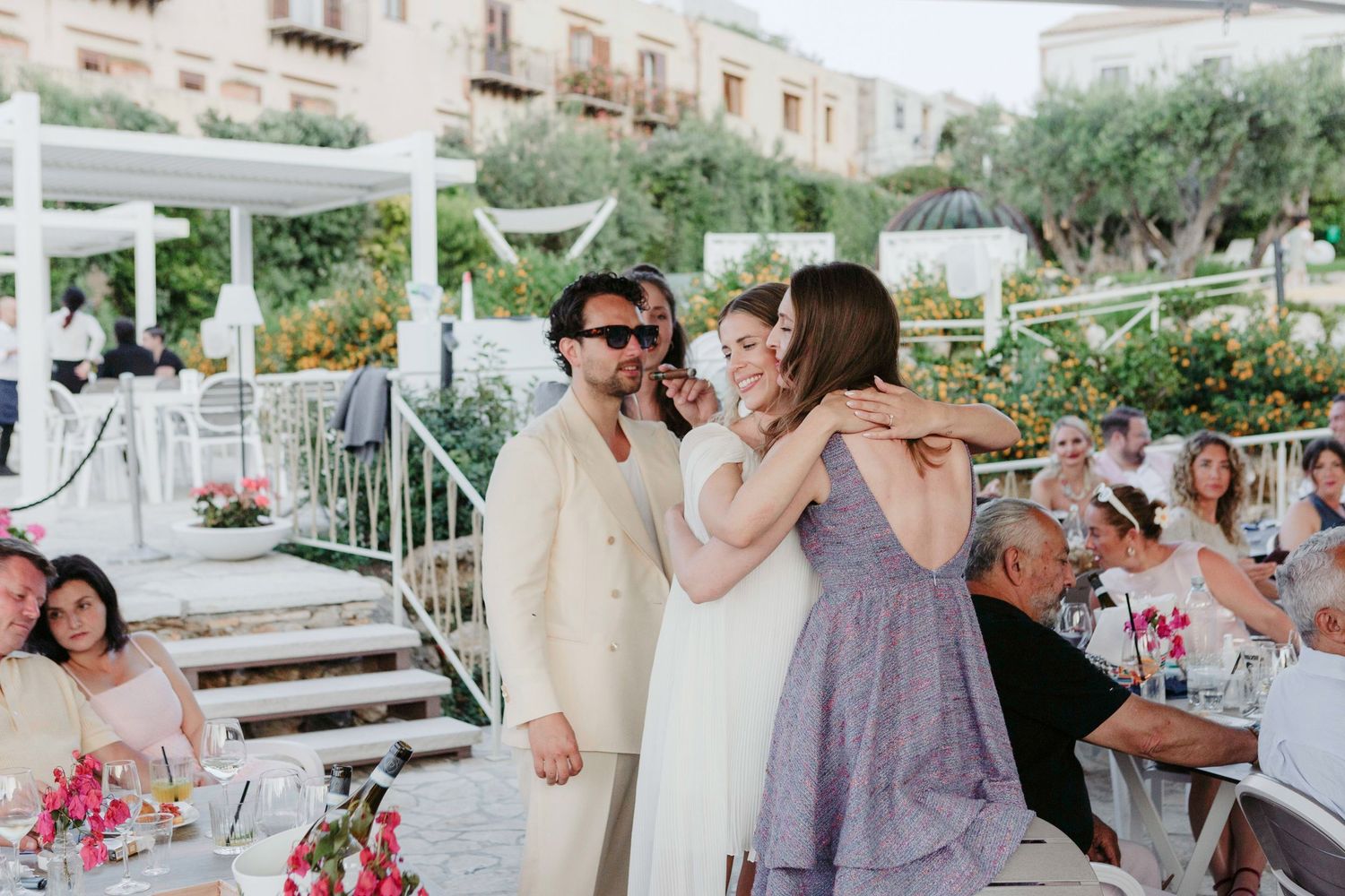 Wedding guests gather outdoors at a scenic venue with string lights and pink floral arrangements during a summer celebration.