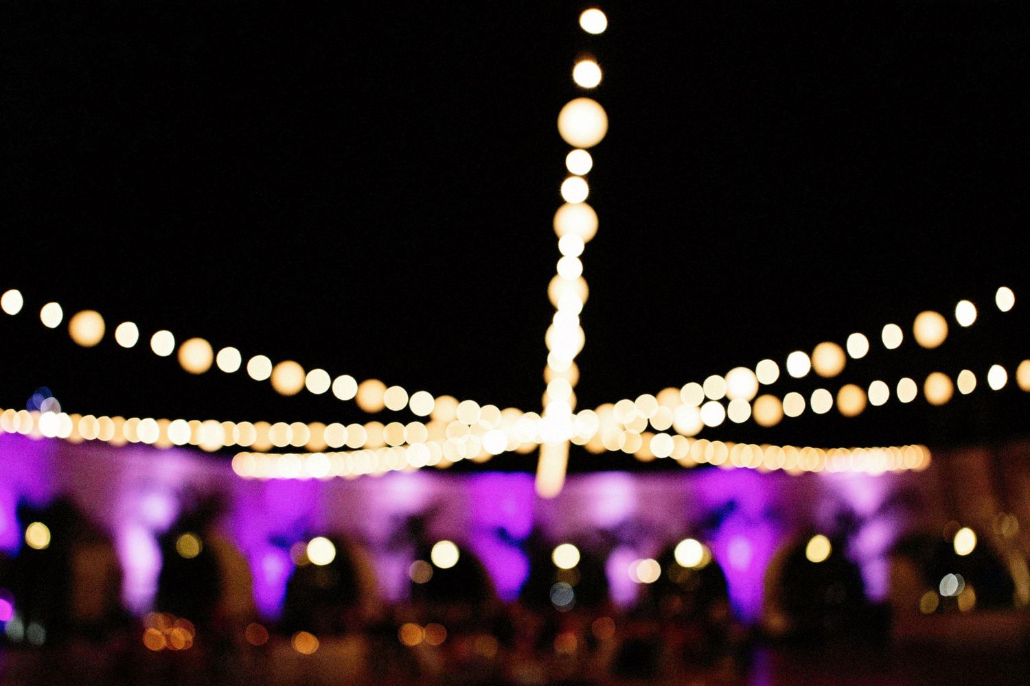 Blurred bokeh lights at an outdoor night event with glowing string lights and purple ambient lighting.