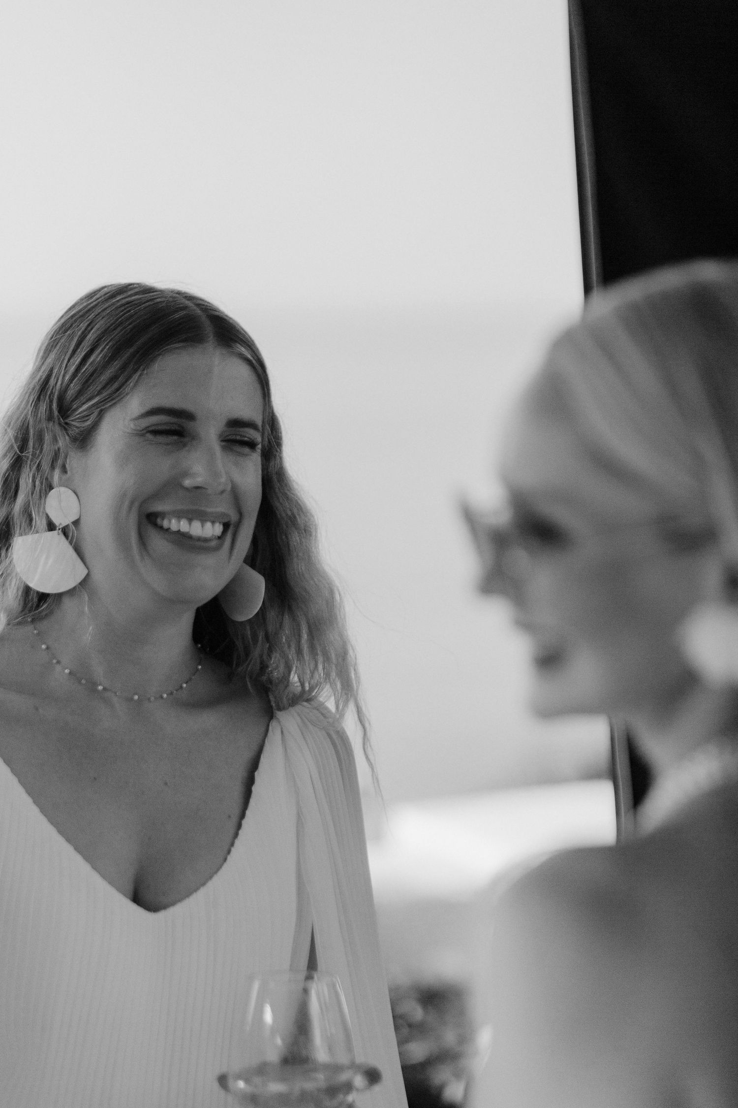 Two women share a candid moment at an event, with one wearing white and statement earrings in black and white photography.