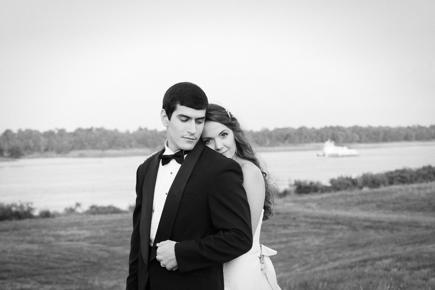 Black and white romantic portrait of a couple in formal attire embracing by the waterfront with a sailboat in the distance.