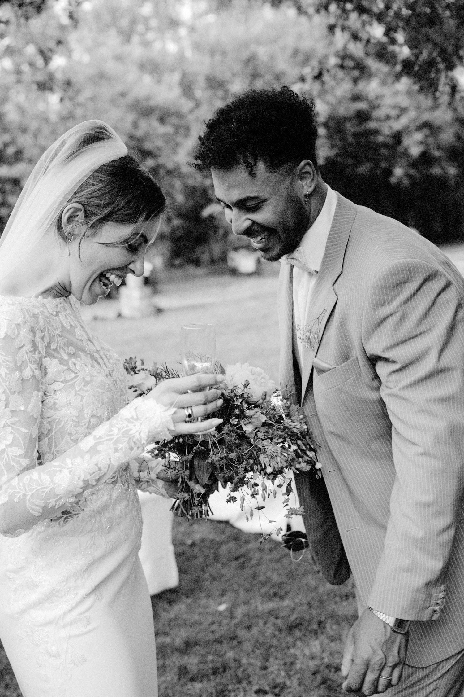 Black and white photo of a laughing bride and groom sharing a joyful moment with a bouquet on their wedding day.