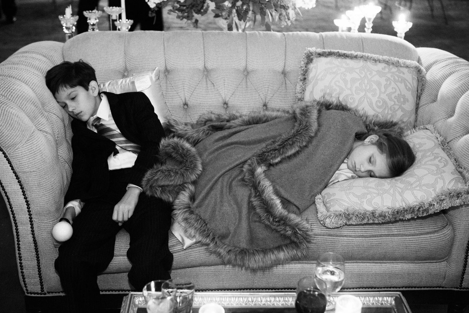 Black and white photo of two children on a sofa, boy in suit and girl lying down in fur-trimmed coat at formal event.