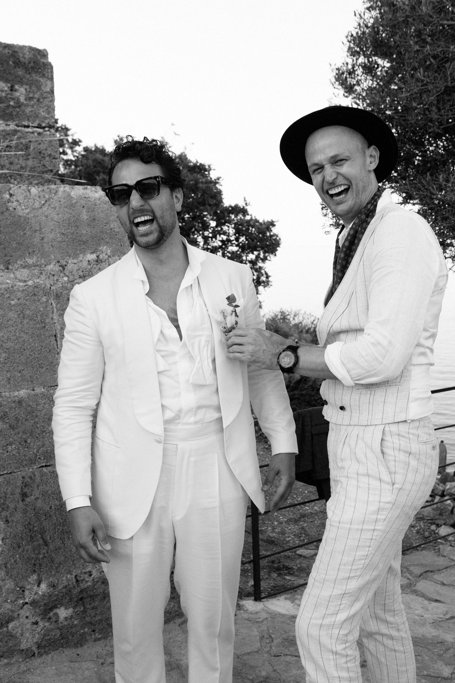 Two laughing men in stylish suits, one in white with sunglasses, pose outdoors in a black and white photo.