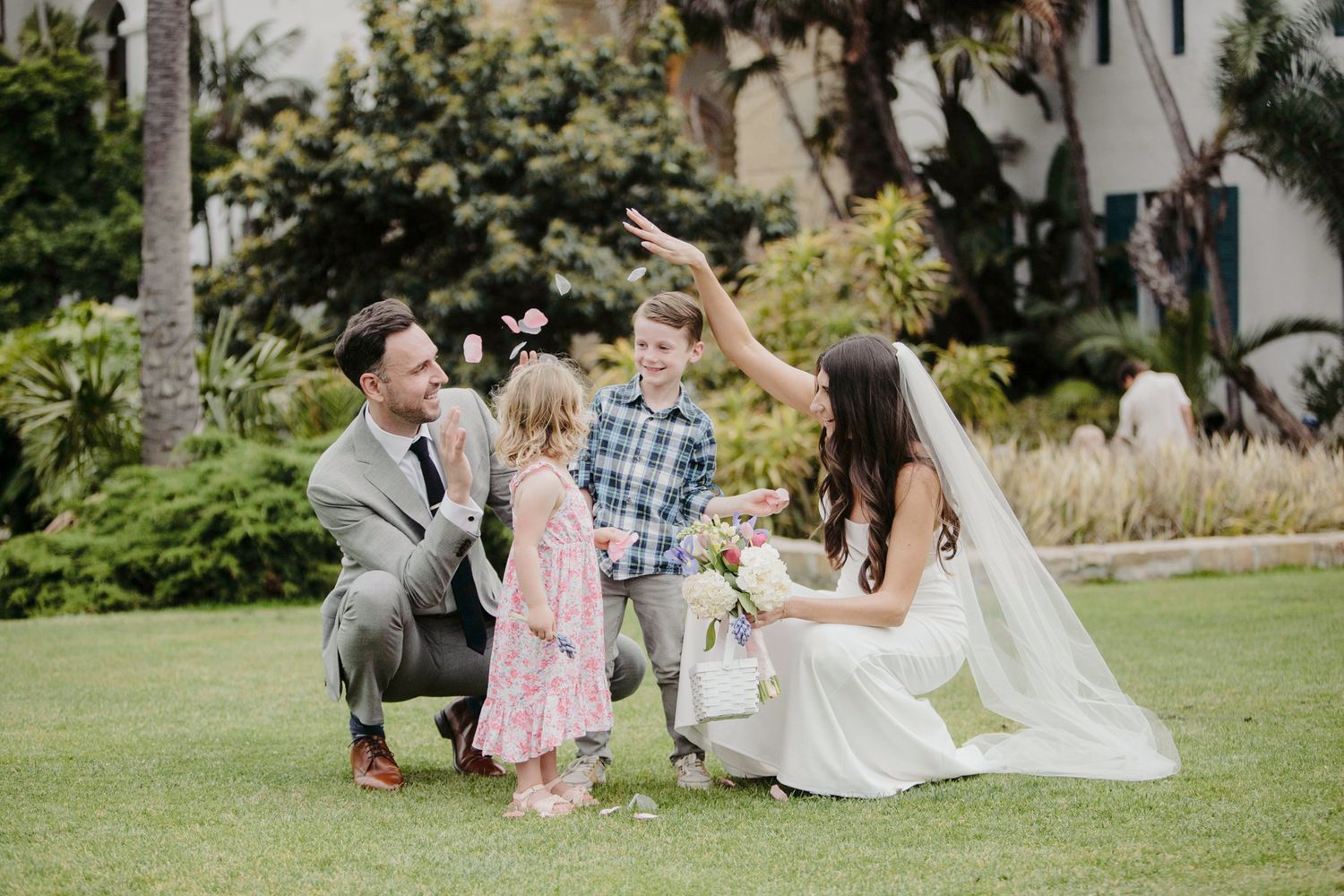 Wedding party enjoying a relaxed outdoor celebration on a lush green lawn with palm trees in the background.