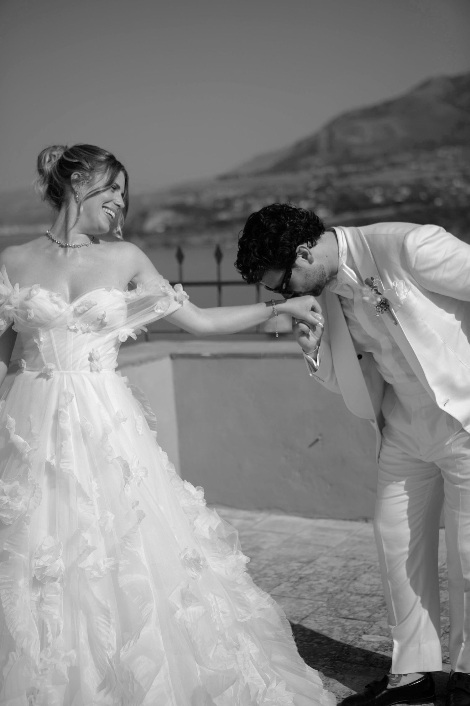 A romantic black and white wedding photo showing a couple in white attire with the groom bowing to kiss the bride's hand.
