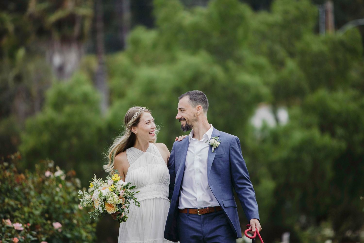 A couple in wedding attire walks together outdoors, surrounded by lush greenery while sharing a joyful moment.