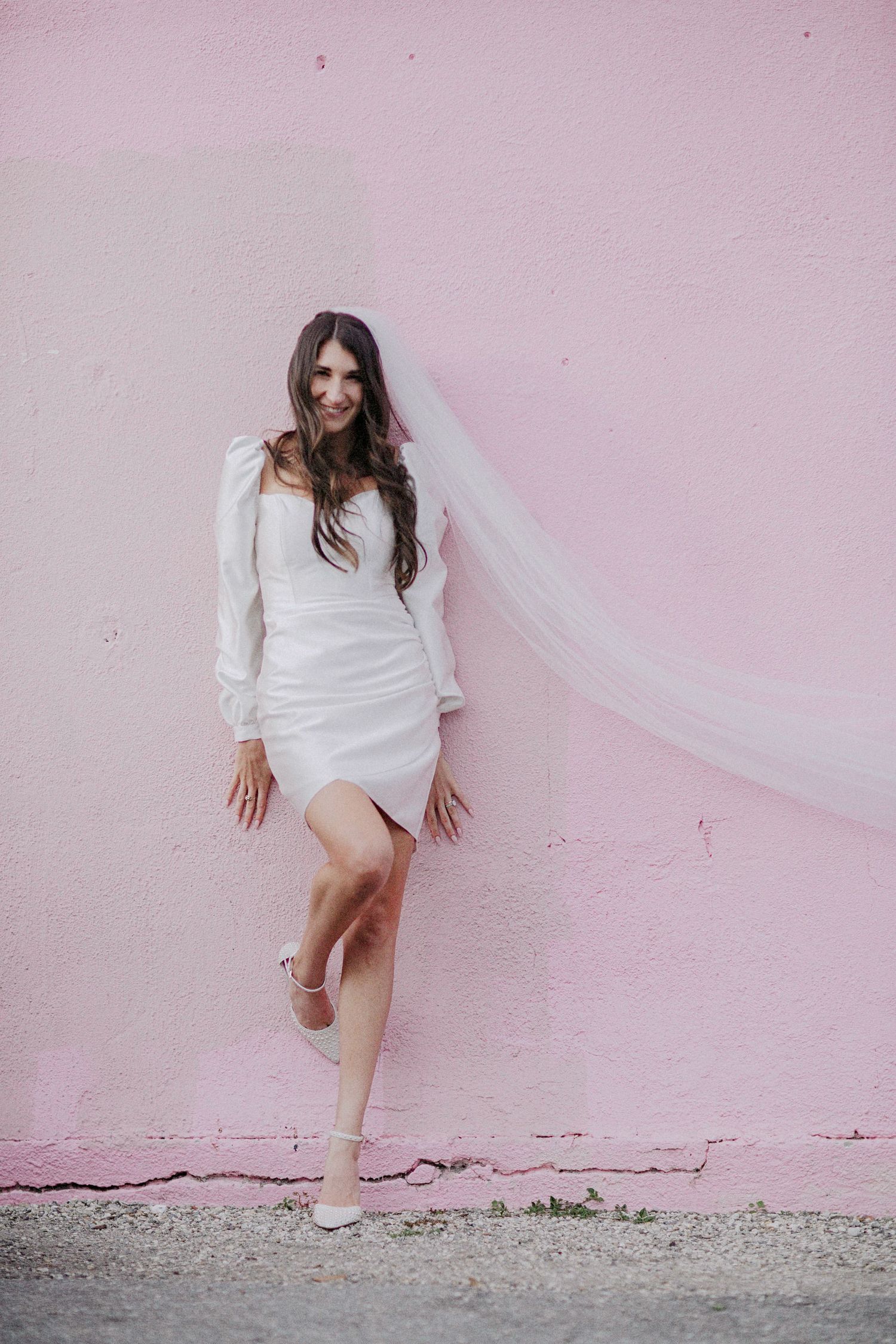 A bride in a short white dress and veil poses against a pink wall, creating a modern minimalist bridal look.