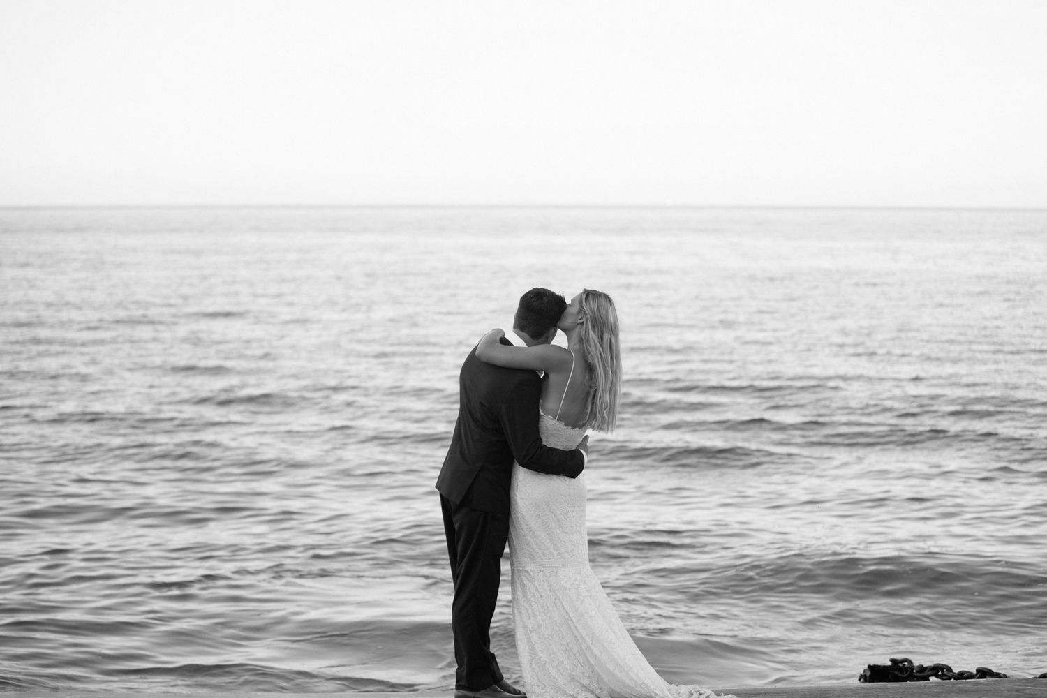 Black and white photo of a couple embracing on a beach in wedding attire with ocean waves in the background.