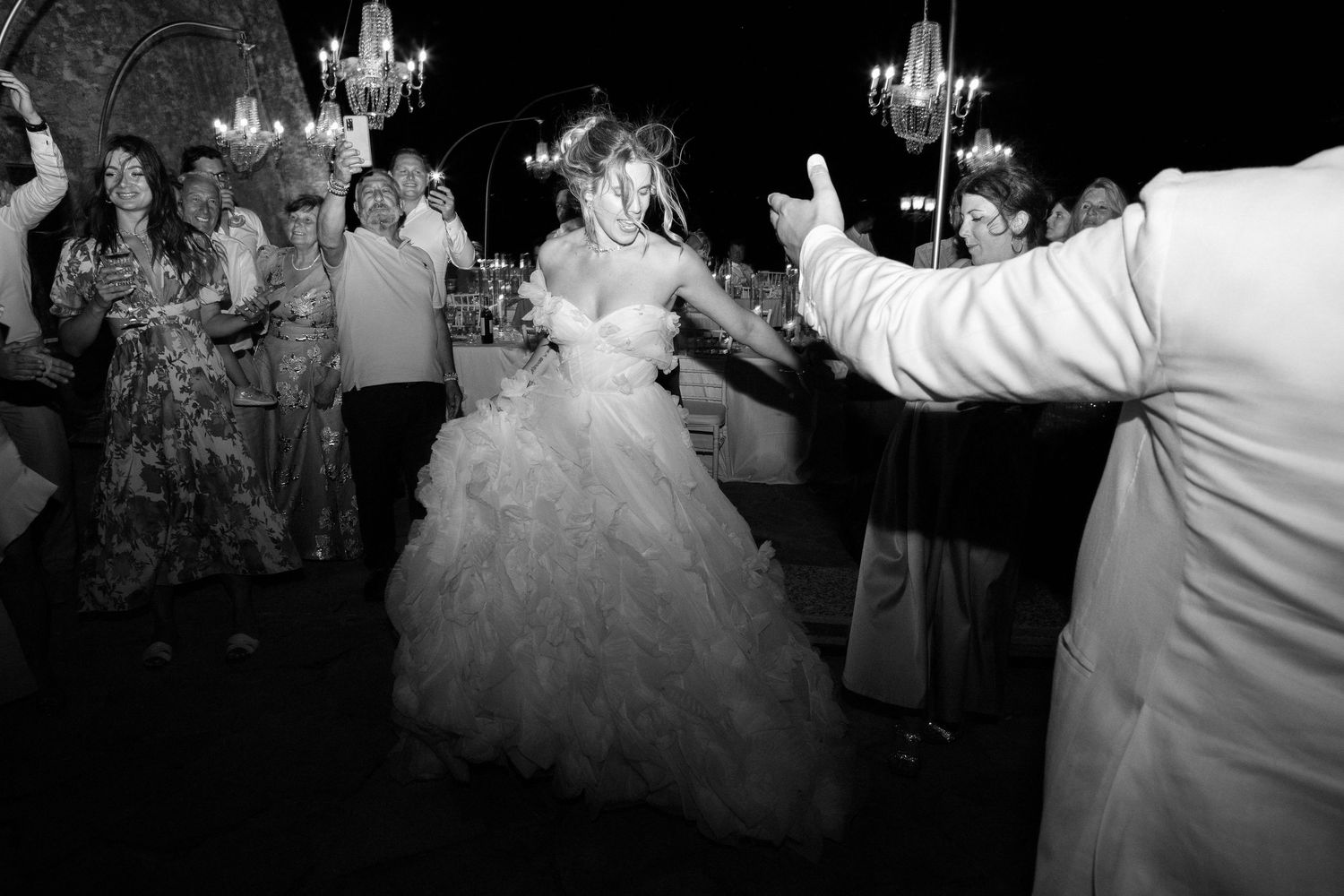 Wedding guests dance and celebrate on a crowded dance floor under elegant chandeliers at a reception party.