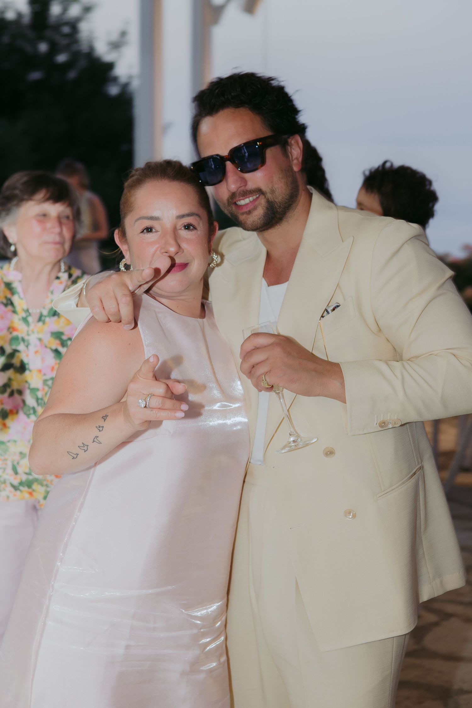 Couple in white attire posing at outdoor wedding reception, woman holding cigar, man in cream suit with sunglasses holding martini.