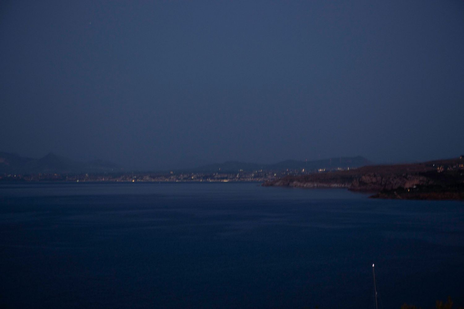 Dark blue waters of a coastal bay at dusk with distant shoreline silhouettes and a single light visible.