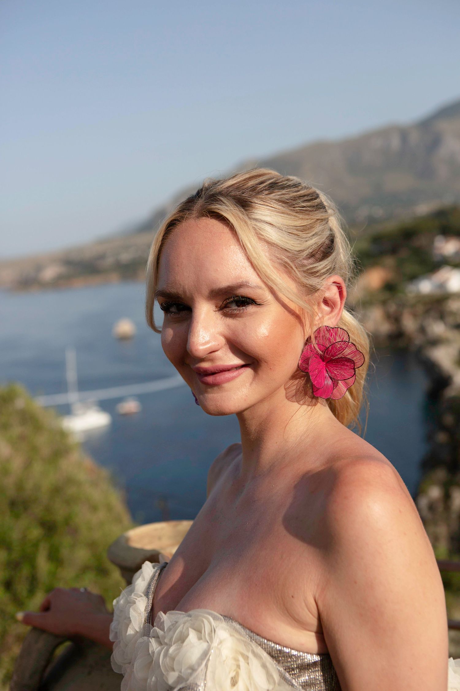 A bride in a white dress with a pink flower in her hair poses against a coastal mountain landscape background.