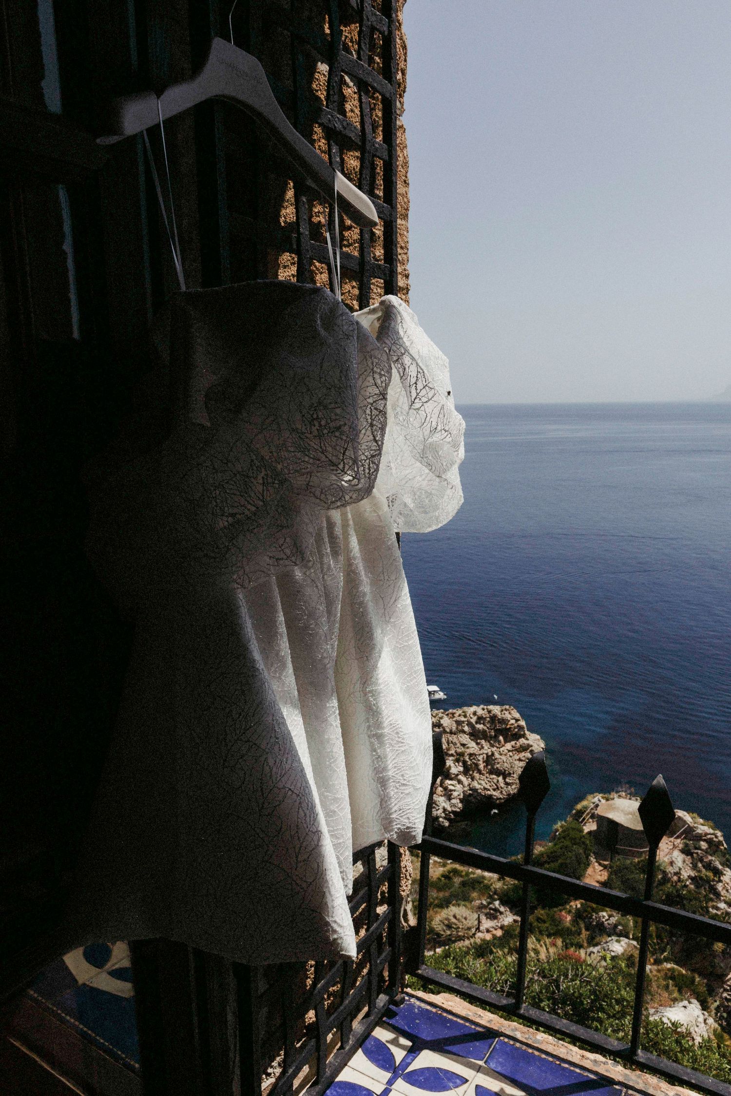 A white dress hanging on a balcony overlooking the Mediterranean Sea with rocky cliffs and blue waters below.