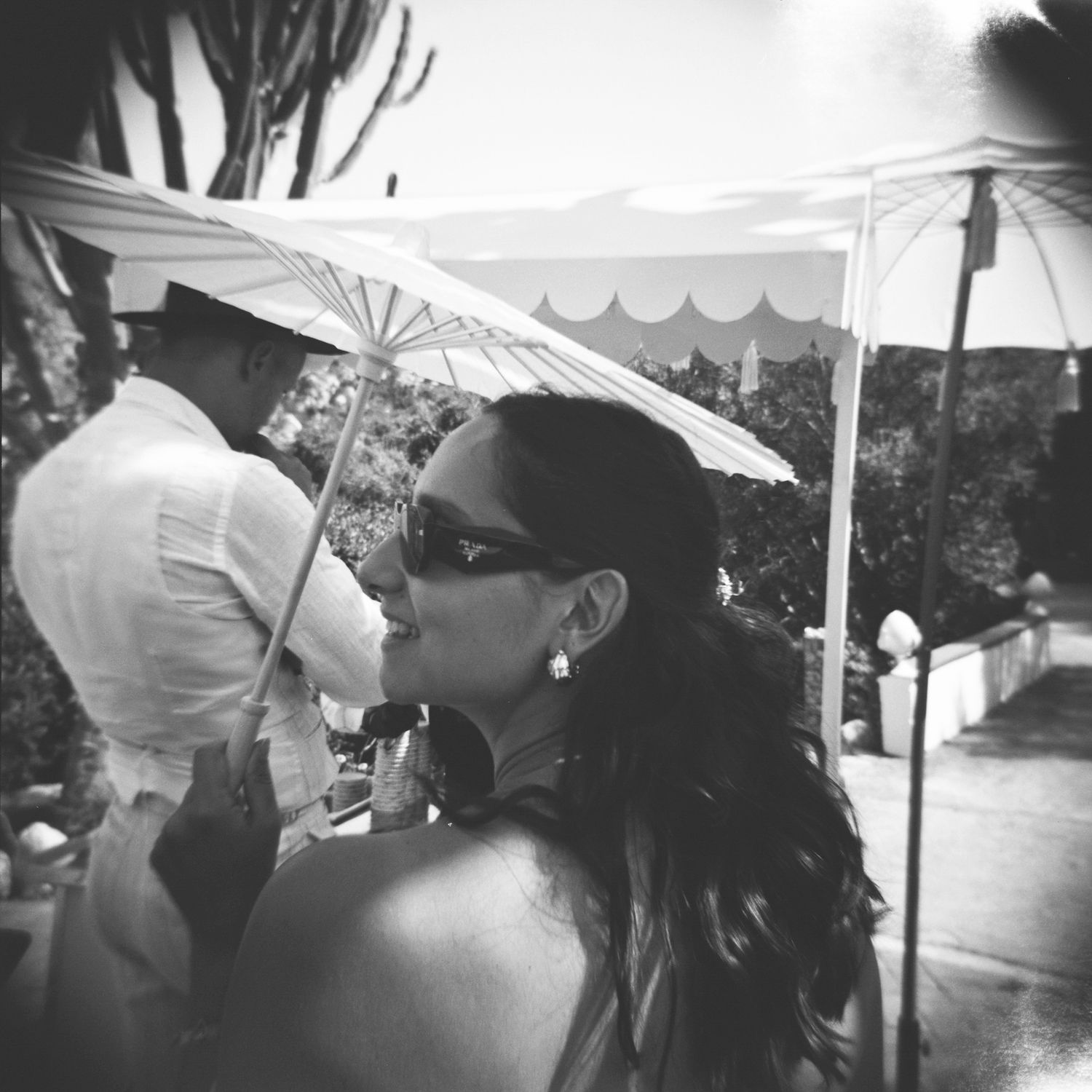 A person in sunglasses holds a white parasol at an outdoor event in black and white vintage style photography.