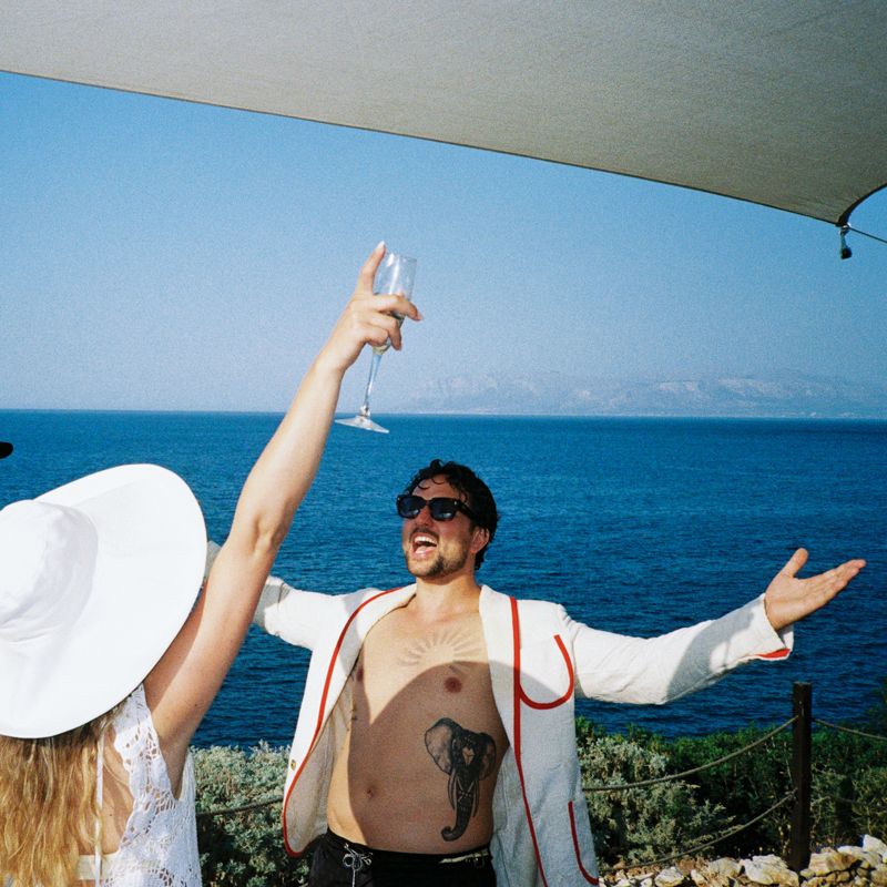 Man in open white jacket raises champagne glass outdoors, overlooking blue sea, with woman in white hat beside him.