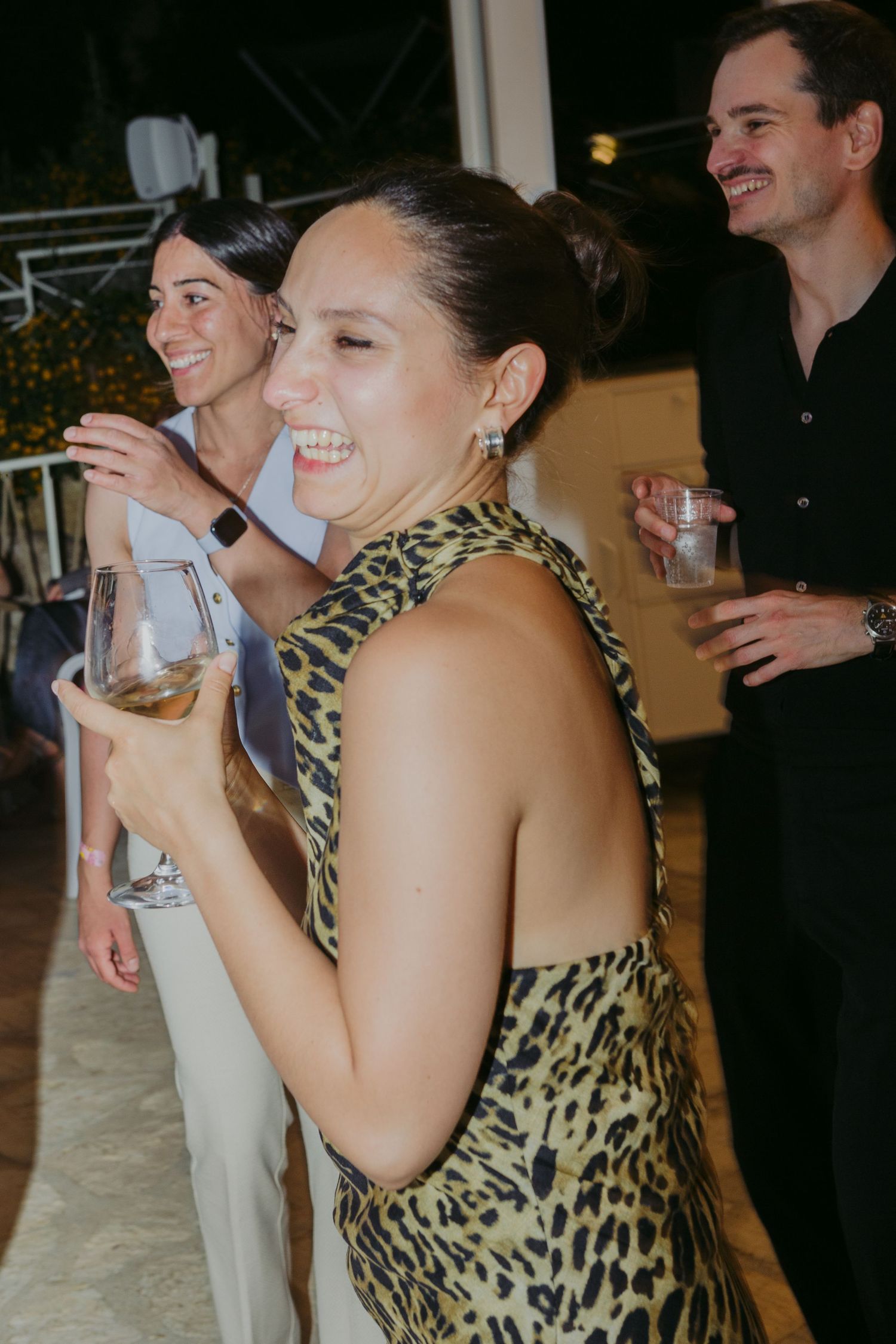 Woman in leopard print dress laughing at evening party, holding wine glass with guests socializing in background.