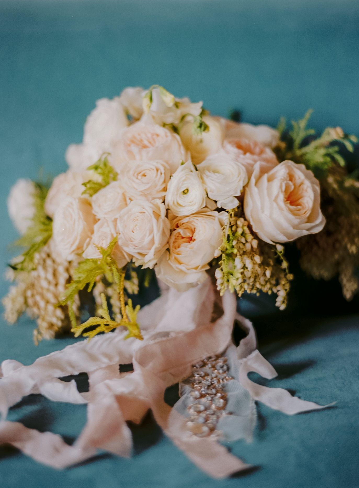 Elegant bridal bouquet of blush roses and ranunculus tied with silk ribbon and pearl bracelet on teal background.