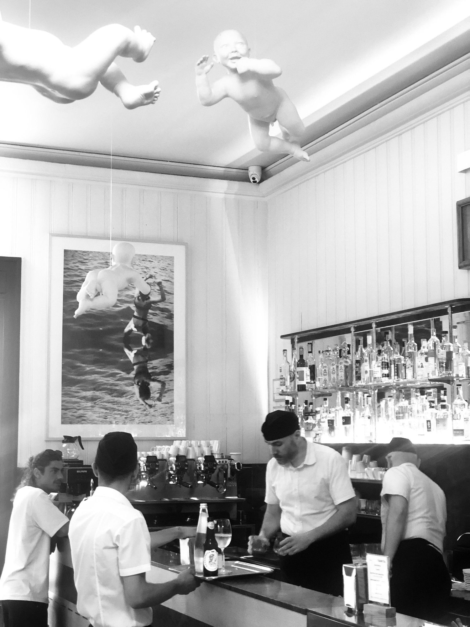 Black and white photo of a bar interior with patrons seated at counter while bartender serves drinks, with artwork on wall.