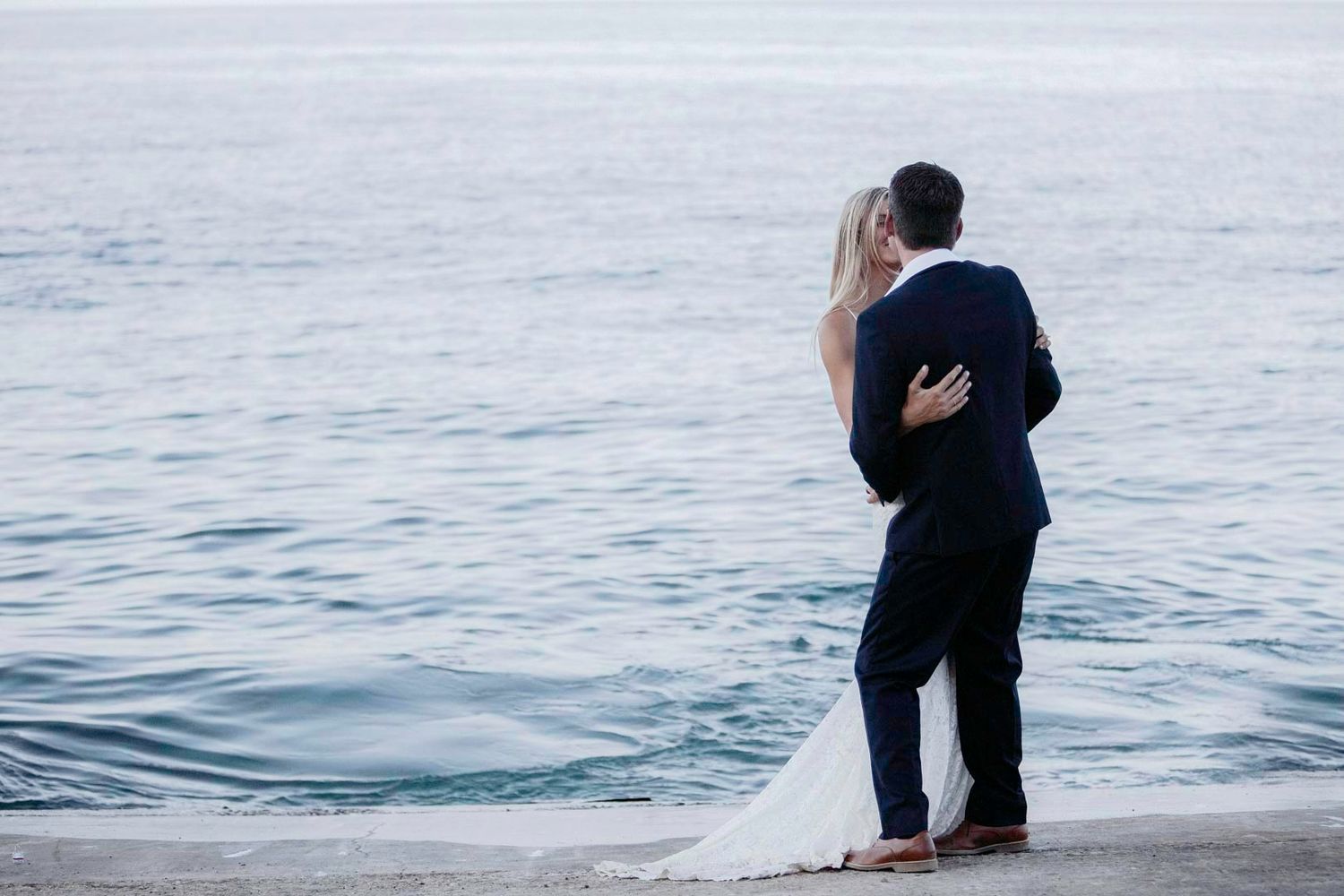 Bride and groom embracing by the ocean shore, gazing at calm blue water during a romantic seaside wedding.