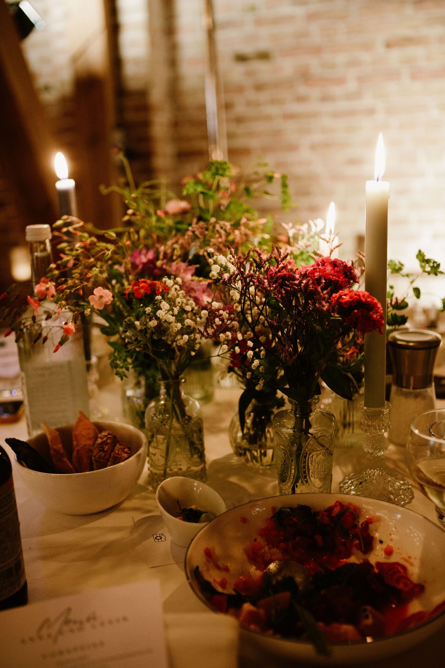 Romantic dinner table setting with lit candles, mason jar flower arrangements, and rustic brick wall backdrop.