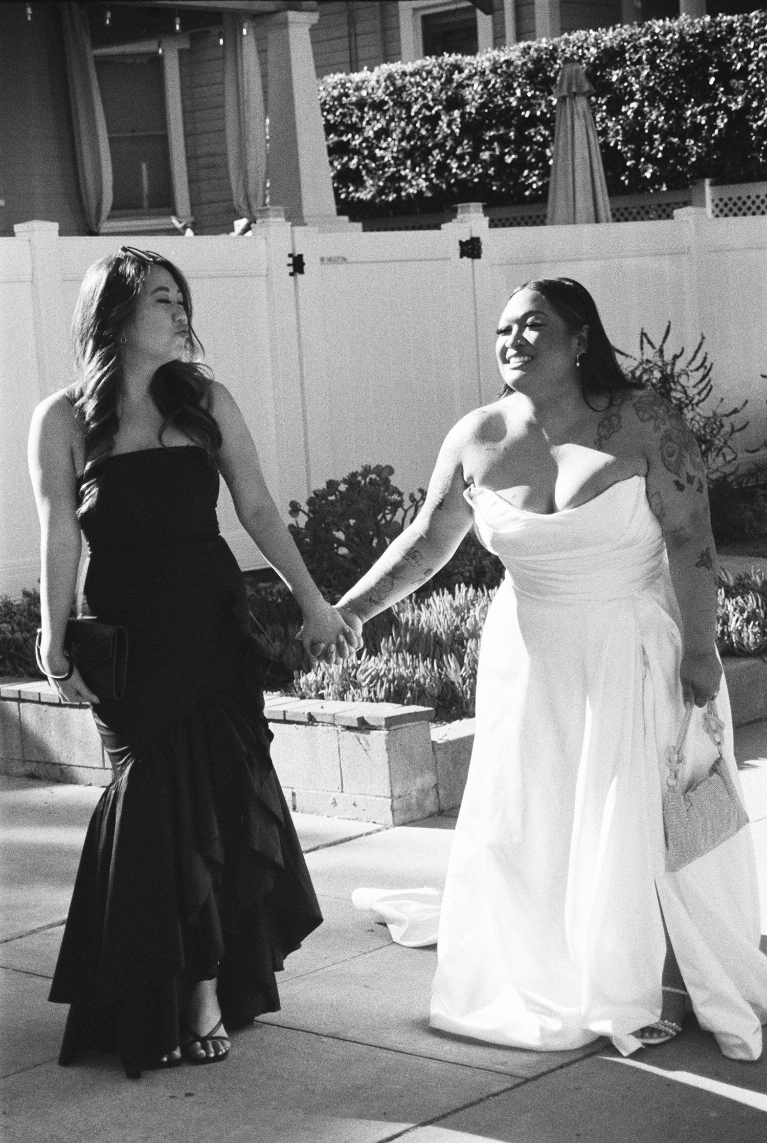 A group in black dresses and white gowns dance and twirl together on a paved walkway in front of a building.