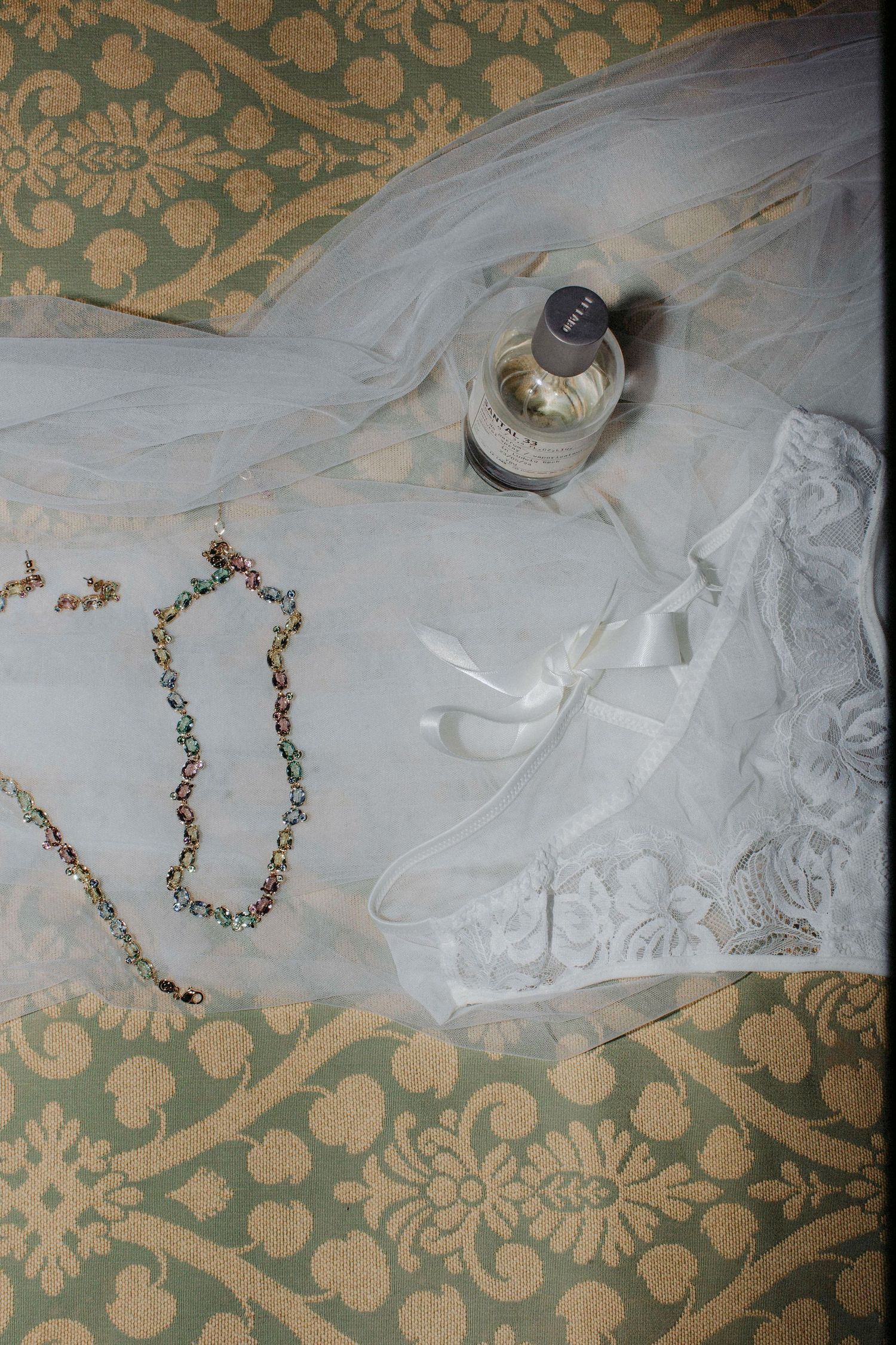Wedding accessories including a white veil, delicate necklace and perfume bottle arranged on a decorative patterned fabric.