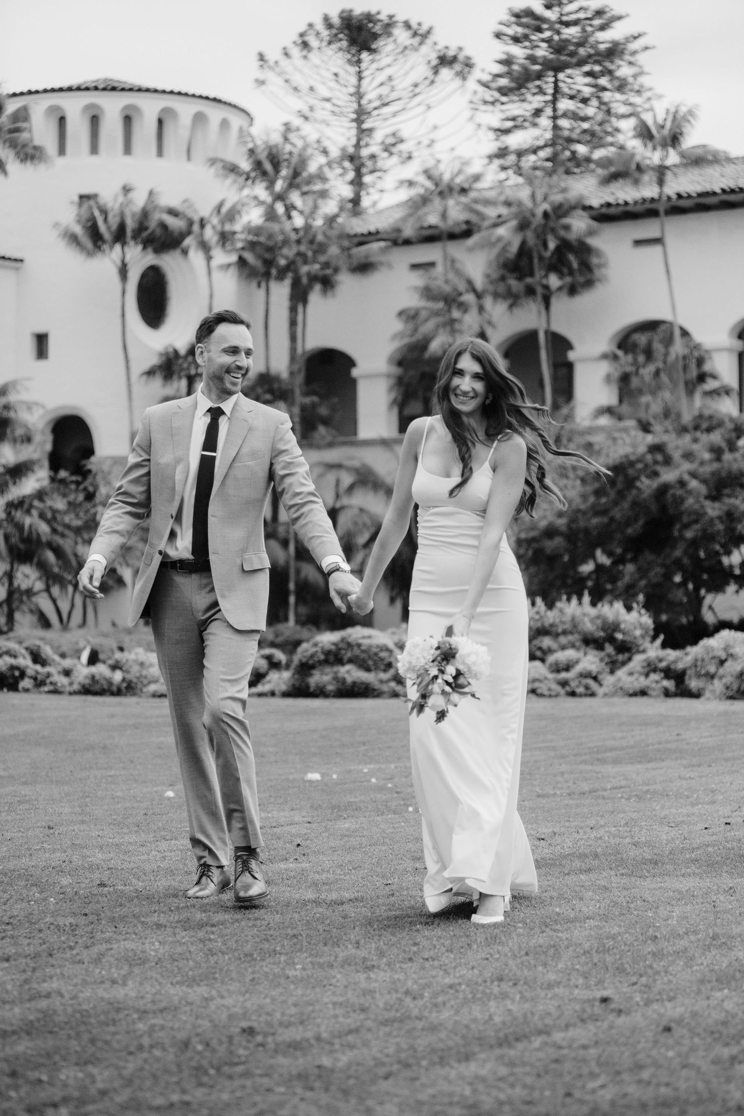 Black and white wedding photo of couple walking hand in hand on lawn with Spanish-style building in background.