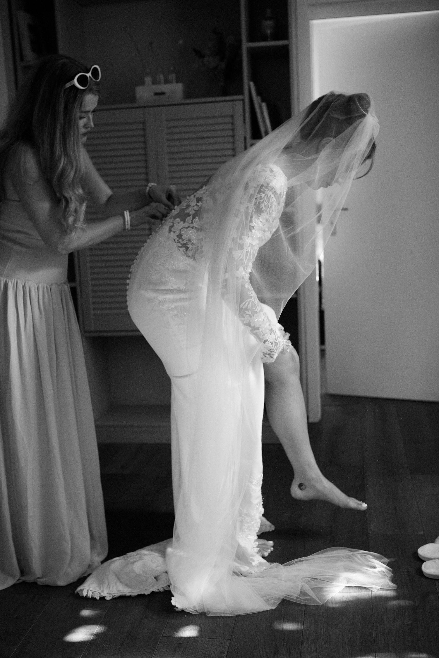 Black and white photo of bride in lace wedding dress and veil being helped by bridesmaid while getting ready.