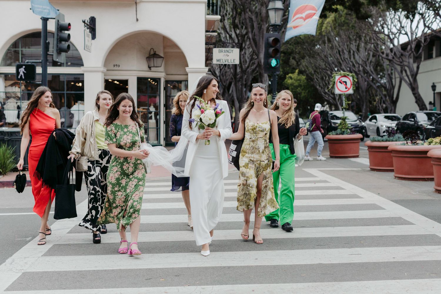 A group of people walking in a bright outdoor plaza with white arches and potted plants in the background.