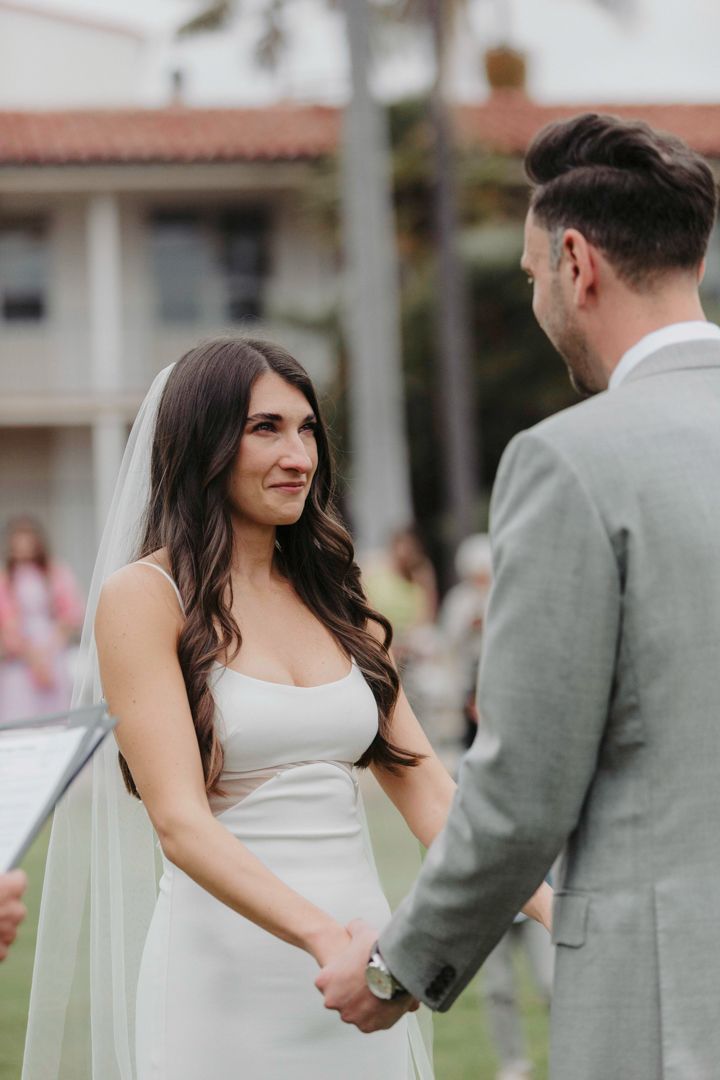 A couple in wedding attire shares a joyful moment on a lawn with Spanish-style buildings in the background.