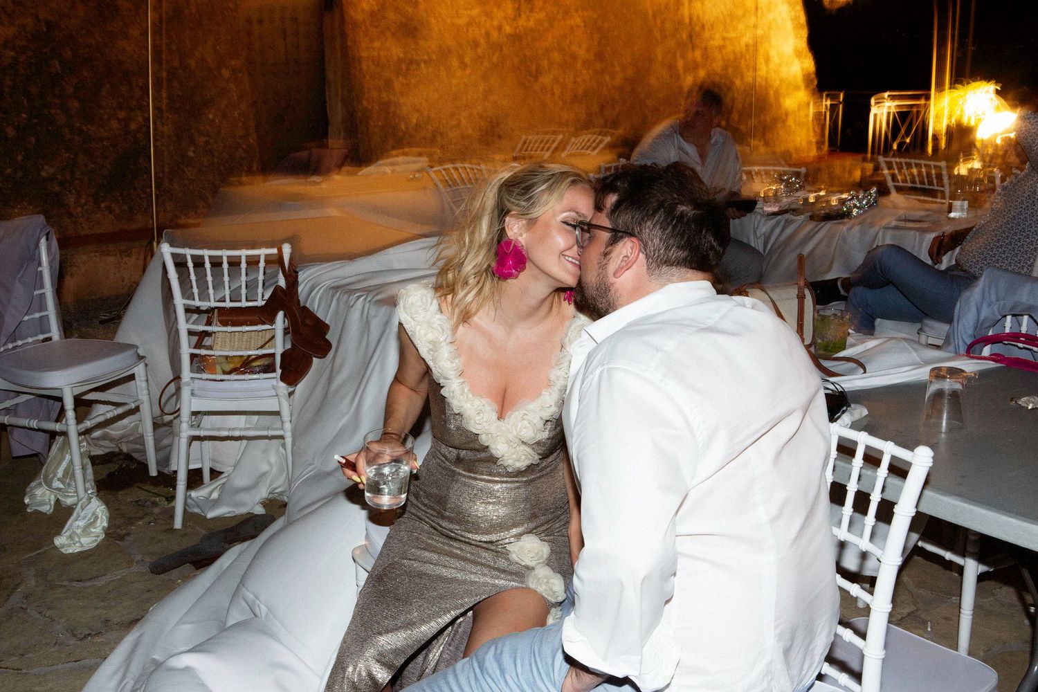 Wedding reception guests dancing and celebrating at an indoor venue with white chairs and decorative lighting.
