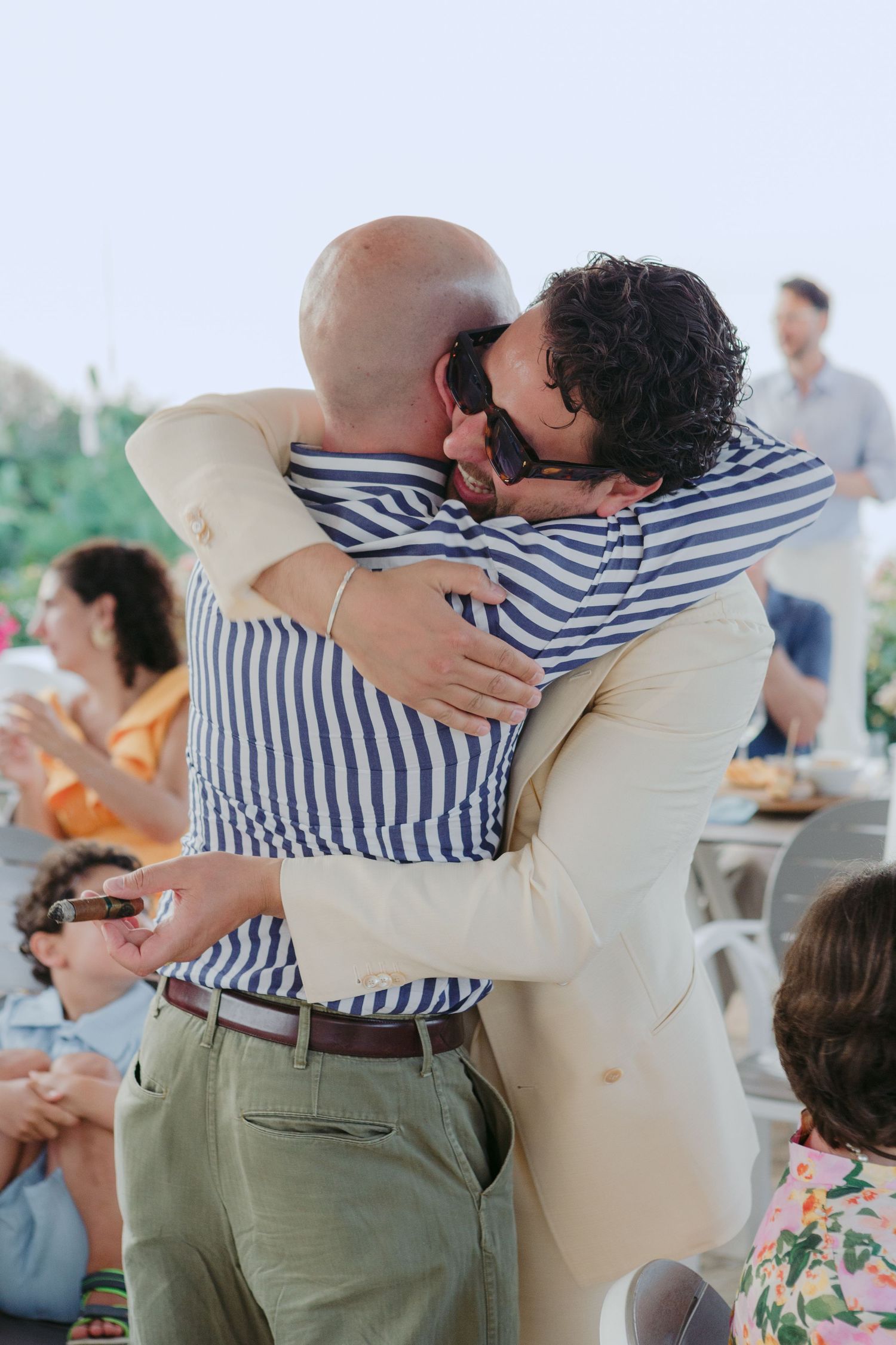 Two people share an emotional embrace at a casual gathering, one wearing a striped shirt and khaki pants.