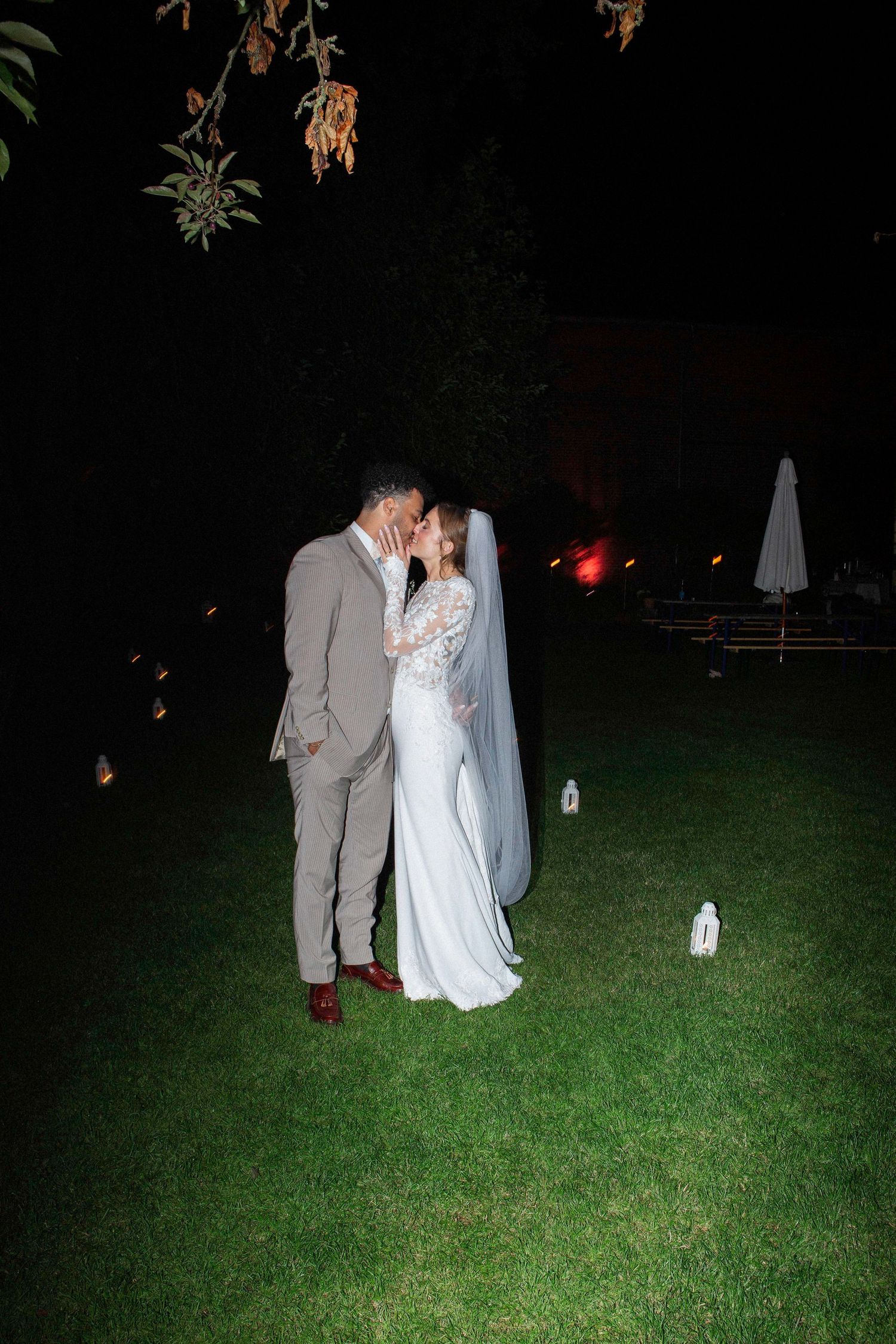 A couple in wedding attire share an intimate moment on a dark lawn illuminated by soft lighting.