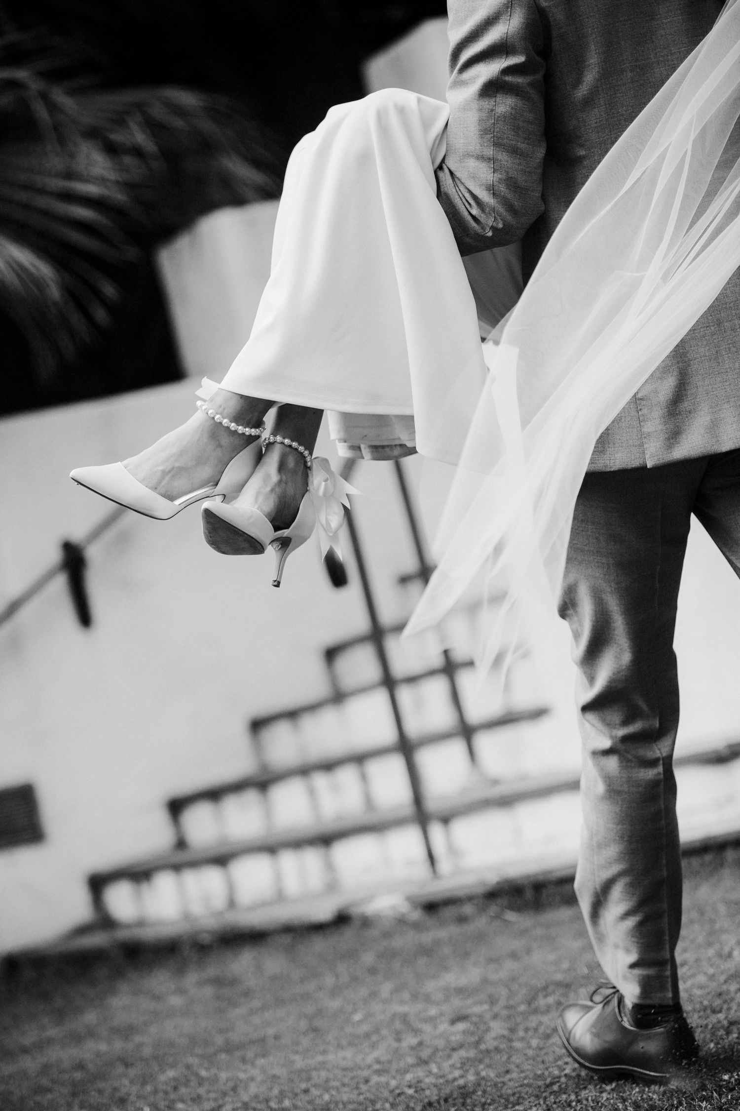 Black and white wedding photo showing elegant high heels and flowing white dress hem against grass.