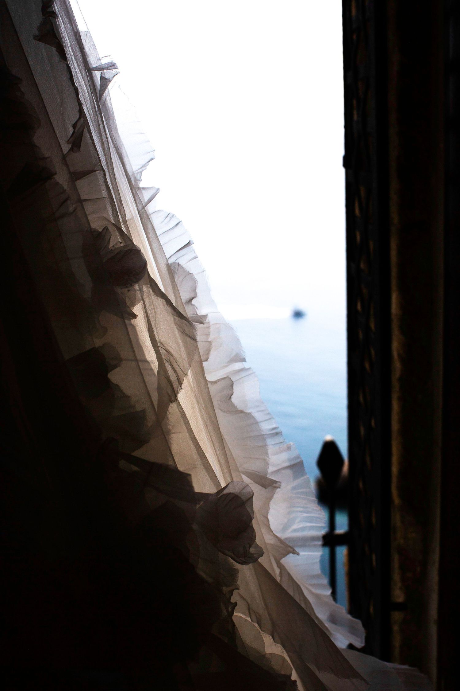 A silhouetted figure stands at the edge of a dark doorway looking out at a bright blue sky through a sheer curtain.