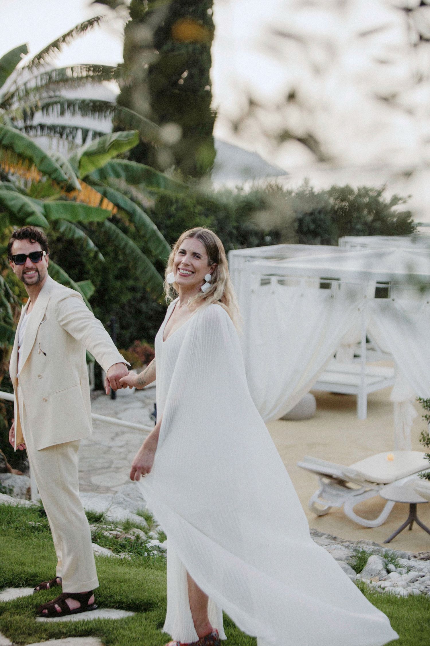 A couple in elegant white attire poses on a lawn with palm trees and lounge chairs in the background.